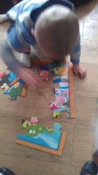 A young child is kneeling on a wooden floor, assembling a colourful Ravensburger Giant Floor Puzzle featuring cartoon animal characters. The child's head is bowed and their hand is placing a puzzle piece.