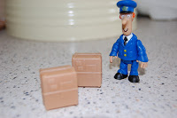 A small toy figure of Postman Pat in a blue uniform stands on a speckled worktop next to two brown parcel-shaped toy boxes, reminiscent of the Classic Vehicle Toy series.