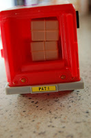 A close-up of a red Classic Vehicle Toy lorry with beige boxes inside. The number plate reads "PAT 1," referencing Postman Pat. The lorry sits on a light-coloured, speckled surface.
