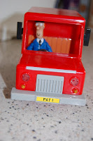 A classic toy red van with a plastic figure of Postman Pat as the driver sits on a speckled worktop. The yellow number plate on the front reads "P4T 1".
