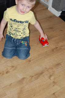 A young child with fair hair, wearing a yellow T-shirt and blue shorts, kneels on a wooden floor while holding a small red Classic Vehicle Toy.