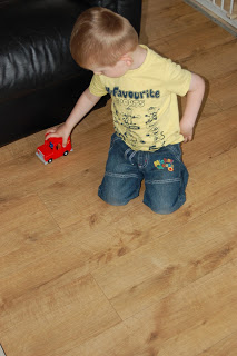 A young child wearing a yellow T-shirt and blue shorts kneels on a wooden floor, playing with a classic vehicle toy lorry near a black sofa.
