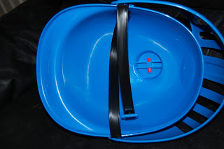 A close-up of a blue plastic bowl with a black strip and two small red clips inside, placed on a dark surface—part of Mike's Mission Set from Mike the Knight.