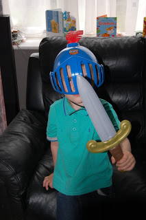 A young child wearing a blue Mike the Knight helmet and holding a plastic sword sits on a black leather chair, with children's books visible in the background—ready for adventure with Mike's Mission Set.
