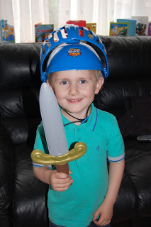 A young boy in a blue polo shirt smiles whilst holding a toy sword from Mike The Knight, wearing a blue plastic helmet, and standing in front of a black sofa with Mike the Knight toys and boxes visible in the background.