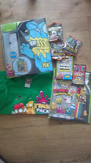 Trash Pack-themed merchandise displayed on a wooden floor, including Trash Pack collectibles like a binder, green T-shirt, trading cards, an activity pad with colouring pencils, and several small packs featuring cartoon-style gross-out characters.