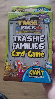 A hand holds a box of "The Trash Pack Trashie Families Card Game," featuring collectable toys and cartoon rubbish characters on the packaging. Suitable for ages 5 and up, for 2-4 players, with giant playing cards.