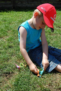 A young child in a red cap, blue sleeveless shirt, and denim shorts sits on grass, excitedly exploring dinosaur figures for a fun Dinosaur Train toy review.