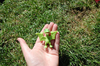 A hand holds a small green toy dragon with outstretched wings, set against a background of green grass in bright sunlight, perfect for a Dinosaur Train toy review setting.