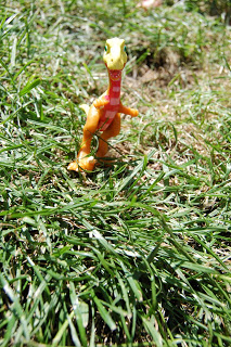 A small, orange toy dinosaur with a pink-striped neck stands upright on green grass in bright sunlight, perfect for any Dinosaur train toy review.
