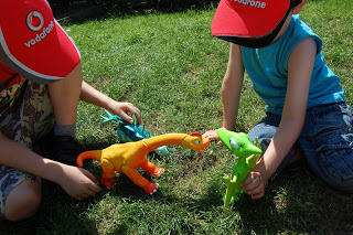 Two children wearing red caps play with colourful dinosaur toys on green grass. Inspired by a Dinosaur Train toy review, one holds a yellow dinosaur whilst the other has a green one, both engaging in imaginative play outdoors.