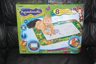 A box of the Tomy Aquadoodle drawing mat features a smiling child lying on their stomach, drawing on the mat. The box highlights “8 Jungle Magic Sounds!” and displays colourful jungle-themed graphics and animals.