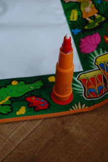 An orange marker stands upright on a colourful Tomy Aquadoodle playmat featuring illustrations of a crocodile, a frog, pink flowers, and drums, placed on a wooden floor.