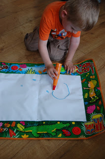 A young child wearing an orange shirt draws with a blue marker on a Tomy Jungle Magic Sounds Aquadoodle drawing mat with a colourful animal border, sitting on a wooden floor.