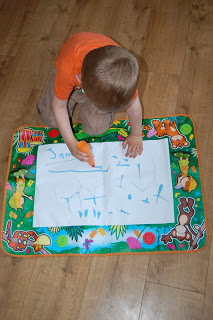 A young child in an orange shirt creates on a large Jungle Magic Aquadoodle mat, drawing with a felt-tip pen while sitting on a colourful, animal-themed play mat spread across the wooden floor.