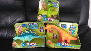 Three toy dinosaurs in green packaging are displayed on a black leather sofa. The toys, perfect for any Dinosaur Train toy review, are labelled as Tiny (green), Morris (blue), and Ned (orange) from the “Dino Ranch” series.
