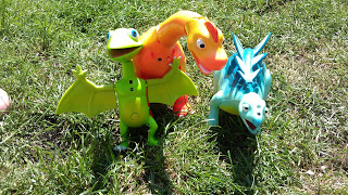 Three colourful toy dinosaurs—one green with wings, one orange with a long neck, and one blue with spikes—stand on green grass in bright sunlight, perfect for any Dinosaur Train toy review or imaginative playtime adventure.