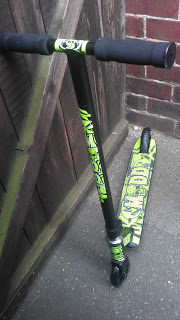 A black and green Madd Gear Beginner Pro Scooter with graphic designs is leaning against a wooden gate beside a brick wall on a concrete surface.