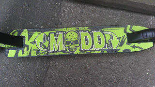 A close-up of a Madd Gear Pro Scooter deck with a green and black design, featuring a skull in the centre and the word "MADD" in bold letters. The scooter rests on a rough concrete surface.