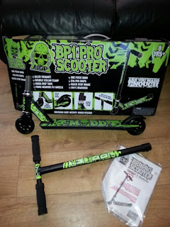 A green and black Madd Gear BP1 Scooter box and partially assembled Beginner Pro Scooter are on a wooden floor. The handlebar, manuals, and assembly tools are placed next to the scooter.