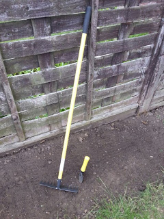 A yellow-handled rake and a small Wilkinsons hand trowel are placed on soil next to a weathered wooden fence in a gardening scene.