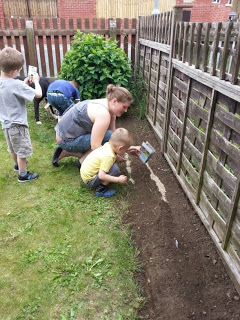 A woman and a young boy kneel beside a garden bed, using gardening tools to dig in the soil near a wooden fence. Two other children stand nearby, one holding a small Wilkinsons spade, outdoors in a grassy garden.