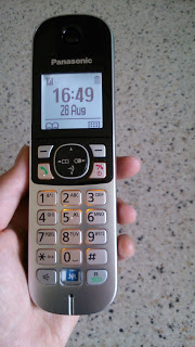 A hand holds a silver and black Panasonic KX-TG6811 digital phone, displaying the time 16:49 and date 26 Aug on its screen, against a speckled worktop background.