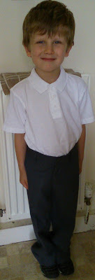 A young boy with short brown hair is smiling whilst standing indoors in front of a radiator, dressed in a white polo shirt and dark trousers—ready for Back to School with new essentials from Asda.
