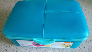 A bright blue plastic lunchbox with a textured lid, divided into three sections, sits on a speckled worktop. Perfect for Back to School season, this Asda school supplies essential features a partially visible product label on the front.