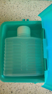 A rectangular, translucent plastic ice pack with a white cap is placed inside an open teal container on a speckled worktop, making it a handy addition to your Asda school supplies for Back to School lunches.