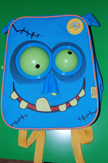 The WOW Pack Wanderer is a blue rucksack designed to look like a cartoon monster face with large bulging eyes, a big smiling mouth with crooked teeth, yellow straps, and decorative stitching. The background is green.