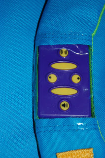 A close-up of a WOW Pack Wanderer purple plastic panel with three yellow buttons featuring icons, sewn into bright blue fabric. The buttons display a speaker, an “O” shape, and a mouth-like shape.