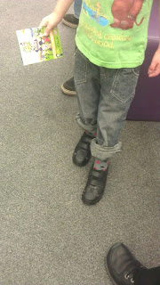 A child wearing rolled-up jeans, a green T-shirt, and black Clarks shoes is holding a colourful card or booklet—perfect for starting school. The photo is taken indoors, showing only the child’s lower half and part of another person’s shoe.