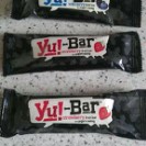Three Yu! Snacks Bar snack bars in black wrappers are placed on a speckled worktop. The labels are visible, with one bar in blueberry flavour and two bars in strawberry flavour—perfect healthy snacks to try for your next snacks review.