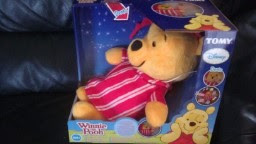 A plush Winnie the Pooh toy wearing red and white striped pyjamas is displayed in a blue Disney-branded box with the TOMY logo, sitting on a dark surface.