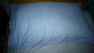 A light blue Slumberdown pillow with a white chequered pattern rests on a white bed sheet. The pillow is positioned against the headboard of a bed, ideal for those seeking comfort, as noted in many Pillow Review articles.