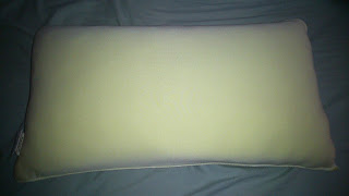 A rectangular, light-coloured Slumberdown pillow is lying on a darker bed sheet. The pillow appears clean and slightly illuminated, with smooth fabric and rounded corners—perfect for anyone seeking a reliable pillow review.