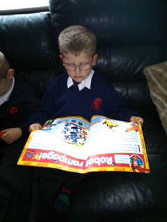 A young boy wearing glasses and a school uniform sits on a dark sofa reading a colourful DC Super Friends magazine called "Robot Rampage!" Another child sits beside him, partially out of frame.