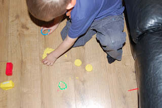 A young child sits on a wooden floor, playing with Skwooshi yellow modelling dough and shape cutters—an engaging Activity Set—next to a black sofa.