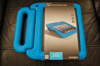 A blue Kensington SafeGrip iPad Case and stand, still in its packaging, is displayed on a black leather surface. The packaging features an image of an iPad in the case and highlights the SafeGrip design for added protection.
