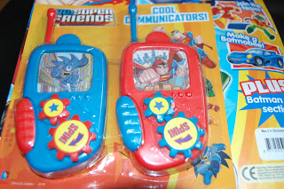 Two toy walkie-talkies themed with DC Super Friends characters, one blue with Batman and one red with Superman, in colourful packaging labelled "Cool Communicators!" and featuring comic-style graphics inspired by DC comics.
