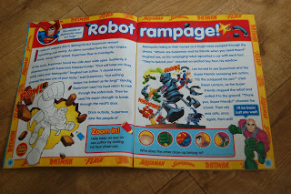 An open children’s book on a wooden floor features colourful comic-style illustrations and bold text titled "Robot Rampage!" with images of Superman from DC Super Friends, a robot, and an unfinished drawing of a character breaking through a wall.