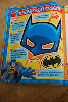 A colourful Superhero magazine page features instructions on how to cut out and wear a blue Batman mask and symbol, with cartoon Batman art and numbered steps in red and white text against a bright yellow and blue background.