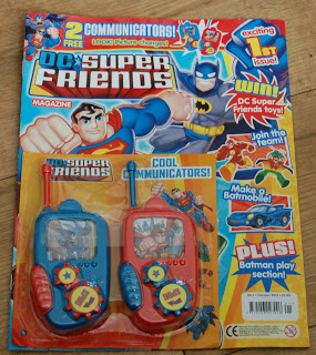 A DC Super Friends Magazine for children, featuring Superman and Batman on the cover, includes two toy communicator walkie-talkies—one red and one blue. Bright, colourful text and comic-style art make this children's magazine eye-catching and fun.