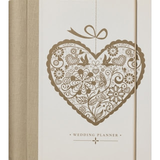 A beige and white Wedding Planner Book with a decorative heart-shaped design featuring flowers, birds, and leaves on the cover, along with the words "Wedding Planner" printed below the heart.