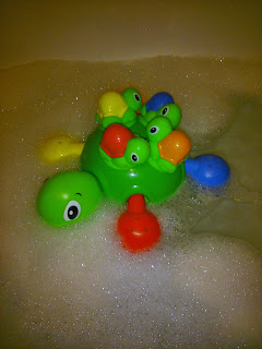 A green plastic turtle from the Tomy Aqua Fun bath toys range carries colourful baby turtles on its back as it floats in soapy bath water surrounded by bubbles.