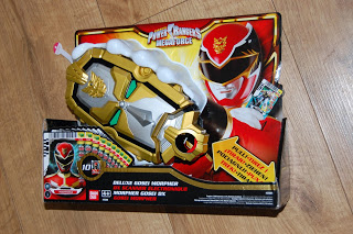 A Power Rangers Megaforce Deluxe Gosei Morpher toy in its packaging, featuring a gold and silver morpher with cards, is displayed on a wooden floor—a must-have for fans and collectors of action-packed toys.