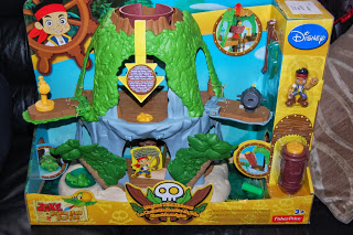 Toy playset for Disney’s "Jake and the Never Land Pirates" features a green and brown plastic island, character figurines, pirate-themed accessories, and a Tiki Hideout in colourful packaging. Fisher-Price and Disney logos are visible.