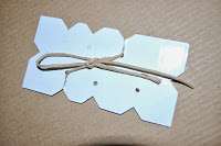 A white card tag with angular edges and two holes is tied with string, placed on a textured brown surface—perfect for labelling treasures at a Tiki Hideout or adventures inspired by Jake and the Never Land Pirates.