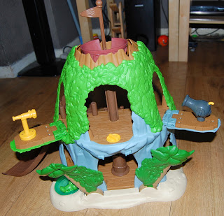 A plastic toy treehouse playset with green foliage, brown platforms, and accessories including a yellow telescope and a grey cannon, inspired by Jake and the Never Land Pirates’ Tiki Hideout, set on a wooden floor indoors.